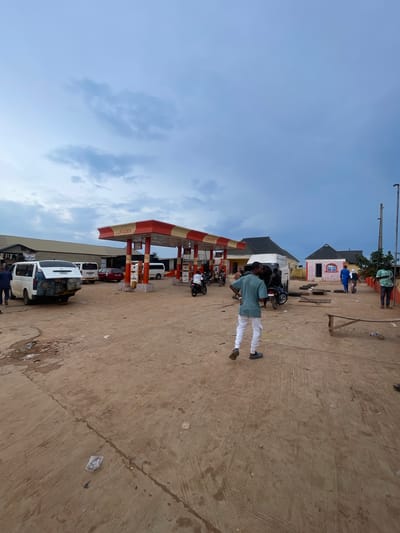 The image depicts a gas station in Osu, Nigeria. In the foreground, a person wearing a green shirt and white pants walks away from the camera. The ground is dusty and uneven, with some debris scattered around.
The gas station features a red and white awning with the name "LUKADEK" visible. Several vehicles are present, including a white van with luggage strapped to its back, a red car, and various motorcycles. Some people are standing around the gas station, while others are on motorcycles. Behind the gas station, there are buildings with corrugated metal roofs.
The sky is overcast with clouds, suggesting it may be evening or a cloudy day. The overall scene appears to be a busy, functional gas station in a rural or semi-urban area of Nigeria.