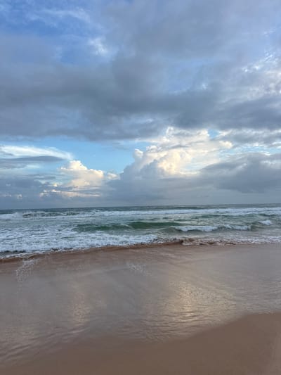 The image depicts a beach scene in Koggala, Sri Lanka. The photograph is oriented vertically, presenting a view of the ocean on the left and the sandy beach on the right. The sea is active with waves, their white foam cresting as they approach the shore. The water appears a murky green, reflecting the overcast sky. The beach itself is composed of wet sand, showing ripples and textures from the receding tide and the light. The sky is filled with dramatic clouds, ranging from dark, heavy storm clouds to brighter, more wispy formations. Patches of blue sky are visible between the clouds, and some sunlight breaks through, illuminating parts of the cloudscape and casting a sheen on the wet sand. The overall mood is serene and somewhat dramatic, perhaps suggesting the approach or aftermath of a storm. The time of day is not precisely indicated but appears to be either early morning or late afternoon, given the diffused light. There is no visible text in the image. The scene is devoid of any people or specific objects, focusing solely on the natural elements of the beach, sea, and sky.