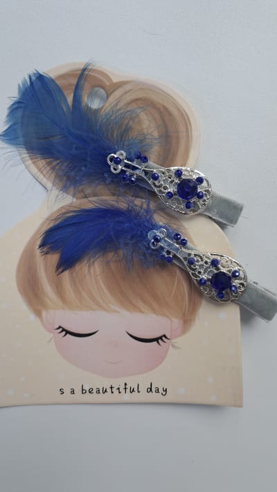 This is a close-up, overhead shot of two blue feather hair clips with ornate silver settings and blue bead embellishments. The hair clips are laid out on a beige card featuring a stylized illustration of a girl's head with closed eyes and blonde hair. The text "s a beautiful day" is printed below the illustration. The background is a neutral, light gray surface. There is no indication of the city of Altai, Kazakhstan within the image.