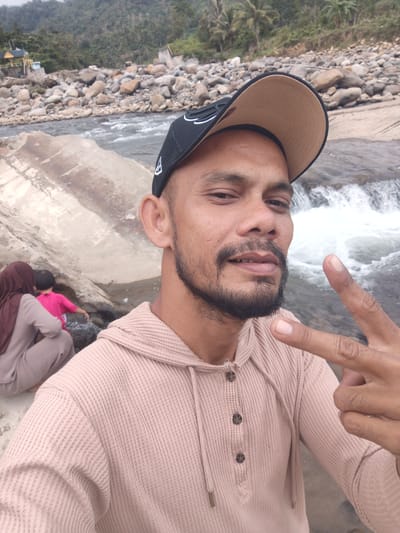 An adult male in Meurah, Indonesia, takes a selfie by a river. He has dark skin, a short beard, and wears a black baseball cap with a tan brim and a light pink waffle-knit hooded henley shirt. His right hand is raised, forming a "peace" sign. In the mid-ground, a woman wearing a reddish-brown hijab and a light brown outfit is seated near large rocks on the riverbank, facing away from the camera. Beside her, a child in a bright pink shirt is near the water. The background features a river with visible whitewater, a rocky shore, and dense green mountains or hills with some scattered palm trees and small structures. The sky appears overcast.