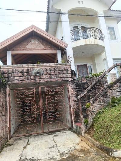 An upward-angled view captures the front facade of "VILLA DAKIRA," a multi-story, light-colored residence in Pô Kô, Vietnam. The property features a secured entrance at the lower level, consisting of closed, ornate reddish-brown wrought-iron gates with scrollwork designs, fastened by a padlock. Above these gates, a stone-block retaining wall displays a central lion's head relief sculpture. Over this wall, a covered entrance area with a gabled wooden roof and white supporting columns bears the embossed text "VILLA DAKIRA" flanked by two bird-like figures. The main building above presents an arched second-story balcony with a decorative black wrought-iron railing and a central white door, flanked by windows. Stains are visible on the wall directly beneath the balcony. A concrete driveway slopes downward from the gate towards the foreground, showing cracks and discoloration. To the right of the driveway, a dark, textured retaining wall with horizontal features supports a grassy embankment with scattered vegetation. No individuals are present in the scene.