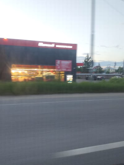A blurry, horizontal, eye-level exterior shot captured from a moving vehicle shows a commercial building on the left side of the frame. The building has a dark facade with a red accent strip along the top edge, and a brightly lit interior is visible through its large windows. A red sign with white lettering is affixed to the facade. In front of the building is a line of green bushes, and behind that, a row of parked vehicles including a motorcycle, a dark SUV, and a lighter colored hatchback. Further in the background, houses with red roofs and green trees are visible, along with power lines and utility poles. A paved road with a white line marking occupies the foreground of the image. The sky is bright, suggesting it is daytime. This image depicts the area near the intersection of Moskovskaya Street and Lunacharskogo Street in Vyalki, Russia.