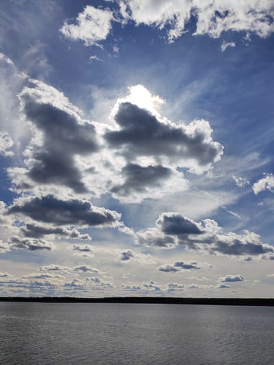 The image shows a body of water, possibly a lake or river, with a treeline visible on the horizon. The sky dominates the view, filled with a mix of white and dark gray clouds against a blue sky. The sun is obscured by clouds, creating a bright glow around them. The water's surface appears calm, reflecting the sky. The image is possibly taken in the vicinity of Topornya, Russia.