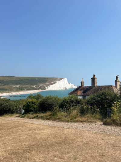 This image shows a view from a hillside towards the Seven Sisters cliffs in Seaford, United Kingdom. In the foreground, dry, tan-colored grass covers the ground, with a gravel path leading through it. A wooden gate is visible on the left. Behind the gate and path, dense green bushes and yellow wildflowers grow. Further back, a small, weathered house with a tiled roof and three chimneys sits nestled amongst trees. Beyond the house, the turquoise sea meets the iconic white chalk cliffs of the Seven Sisters. A beach with a few scattered people is visible at the base of the cliffs. The sky is a clear, bright blue.
