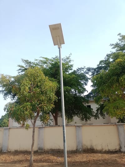 The image depicts an outdoor scene in Panhauya, Nigeria. A tall, silver-colored streetlight dominates the center of the frame. The streetlight has a rectangular white fixture at the top. Behind the streetlight, there are several trees with green foliage. In the background, a beige-colored wall with vertical gray pillars runs horizontally across the scene. Behind the wall, the corner of a white building is visible. The ground is covered with brown dry leaves and dirt. The sky is light blue and clear. There are no people visible in the image. The writing on the streetlight pole says "ABU/AGR/2018-19/SPL".