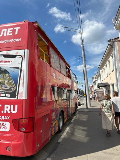 A bright red double-decker bus is parked on a street in a European-style city, likely in Russia given the Cyrillic text on the bus. The bus is adorned with colorful illustrations, possibly depicting local landmarks or cultural motifs. The words "ЛЕТ" and "ЭКСКУРСИИ" are visible on the side of the bus, indicating it is for summer tours or excursions. A portion of a smartphone screen is displayed on the bus, showing an image of a park with a body of water and a ship, alongside text that reads "ОРБИЛЕТ" and mentions discounts. The street is paved with stone, and buildings with light-colored facades and windows line the right side of the road. A utility pole with multiple wires stands near the bus. Two people, a woman and a man, are walking away from the camera on the sidewalk, with the woman wearing a patterned shirt and a long skirt, and the man in a t-shirt and shorts. The sky is blue with scattered white clouds. The overall impression is a sunny day in an urban environment. The location mentioned in the prompt, Putyatino (Semivragovskaya S/A), Russia, is a small village in the Ryazan Oblast, which is unlikely to feature a double-decker tour bus of this scale. Therefore, it is more plausible that the image depicts a tour bus in a larger Russian city.