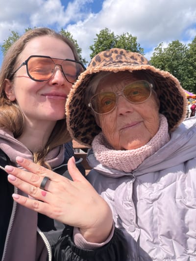 A low-angle selfie shows two people, an older woman and a younger one. The older woman is positioned at the top of the frame and slightly to the right. She wears a leopard-print bucket hat, glasses with reflective lenses, and a light pink knitted collar under a puffy silver jacket. The younger woman is positioned at the bottom of the frame and slightly to the left. She wears glasses with black frames and a pink hoodie under a black jacket. The younger woman's hand is visible on the right side of the frame, with a black ring on the ring finger and a small heart tattoo on the back of her hand. The background shows trees, a partly cloudy sky, and possibly other people in the distance. The location, Valozhyn, Belarus, is not discernible from the image itself.