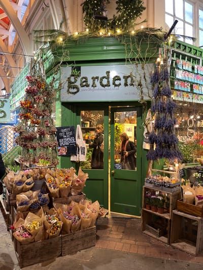 The image captures the exterior of "the Garden" shop, located within the Oxford Covered Market in Oxford, United Kingdom. The shop features a dark green wooden facade with a double entrance door. Above the doorway, a sign constructed from green foliage spells "the Garden," with its website "www.thegardenoxford.co.uk" and a telephone number displayed below. The facade's canopy is decorated with artificial pine garlands and white string lights, from which twisted natural branches and additional string lights descend.
To the left of the entrance, a tall, conical display resembling a tree is formed from bundles of dried pink and orange flowers, wrapped with string lights. Below this, numerous small bouquets of dried mixed flowers, wrapped in brown paper, are arranged in wooden crates on the reddish-brown brick pavement. A black chalkboard sign advertises "CHARITY TOTE BAGS" and displays a small photo of a baby.
To the right of the entrance, a similar conical structure is constructed from bundles of dried lavender, also illuminated by string lights. More bouquets of dried flowers in brown paper and potted plants are displayed in wooden crates on the pavement.
Through the glass panels of the green double door, two individuals are visible inside the brightly lit shop. One person, seen from behind wearing a dark, possibly faux-fur, coat, is inspecting merchandise on a shelf to the left. Another person, wearing a dark coat and a red and black striped scarf, stands further inside the shop. The shop interior appears well-stocked with plants and other items.
The environment is an indoor market setting, evident from the arched, white-framed ceiling structure and overhead lighting visible in the background. The artificial illumination suggests an evening or low-light condition.