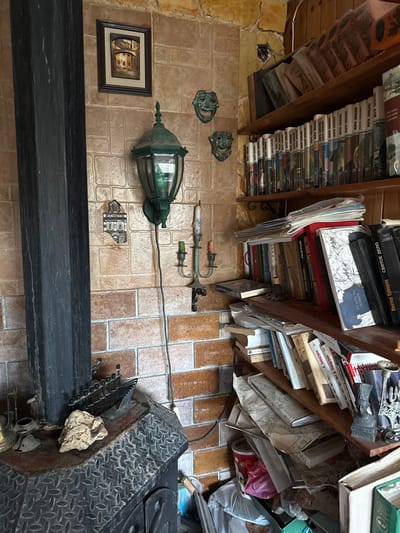 The image depicts an indoor corner of a room located in Binyamina, Israel, characterized by a rustic and densely decorated aesthetic. The walls are primarily covered with rectangular, textured tiles resembling varying shades of brown and tan brickwork, transitioning to rough, light-colored stone or plaster at the upper wall junction.

On the left side of the frame, a tall, dark, square-profile column, likely a stovepipe or structural beam, extends vertically. Below it, a dark-colored, heavy-duty stove or fireplace insert with a diamond plate pattern on its top surface is present. Resting on the stove are a small, dark metal model of a ship, an irregularly shaped piece of light-colored rock or coral, and a small brass container along with other indistinct metallic objects.

Mounted on the tiled wall is a large, dark green, antique-style wall lantern with glass panels, from which a black electrical cord descends. Above the lantern, a rectangular framed picture depicting an architectural scene is affixed. To the right of the lantern, two small, weathered green metallic masks, resembling theatrical tragedy and comedy masks, are wall-mounted. Below the lantern, a small decorative tile depicting a building facade is visible, and further down, an ornate dark metal two-arm candelabra holds two partially consumed, multi-colored candles.

The right side of the image features multiple wooden shelves built into the corner, densely packed with books. The books, many appearing to be hardcovers, display various spine colors, and several visible titles are in Cyrillic script. Books are arranged both vertically and horizontally, interspersed with stacks of papers, maps, and other miscellaneous items. On a lower shelf, a small bust sculpture and an ornate metallic object are visible. The floor area beneath the lowest shelf appears cluttered with items including a red bucket and plastic bags. No individuals are present in the scene.