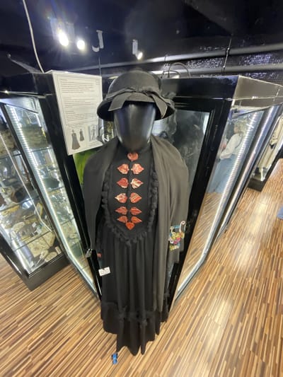 A full-body shot of a mannequin dressed in a dark outfit, including a hat and a long dress with ruffled trim, is presented in a museum setting. The mannequin is positioned in front of a display case, which reflects a person wearing a white top and blue pants. The background features other display cases filled with various artifacts. The flooring is made of wood-look tiles laid in a linear pattern. A sign with text and illustrations is visible behind the mannequin. The overall lighting appears to be from overhead fixtures.