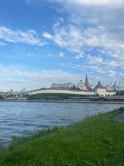 The image shows a wide body of water in the foreground, with rippling waves and a grassy embankment. In the background, across the water, is a complex of buildings, including a white fortress-like wall, several domes of various sizes and colors (some blue, some white), a tall, slender tower with a spiral roof, and other architectural structures. The sky above is a bright blue, with scattered white clouds. The scene appears to be a cityscape viewed from across a river or bay. The location is identified as Дачное, Russia.