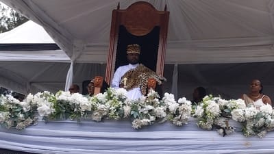 A man wearing a white robe and a leopard skin draped over his shoulders sits on a throne in a large white tent. The throne is made of dark wood and has a carved design on its back. The tent is adorned with white flowers, and there are other people seated in the background, including a woman in white. The location is in Rukoko, Democratic Republic of the Congo, at coordinates latitude -1.624, longitude 29.258. The man is likely a prominent figure in the community, possibly a chief or a king. He is surrounded by other members of his court. The event may be a traditional ceremony or a political gathering. The image evokes a sense of power, tradition, and ceremony.