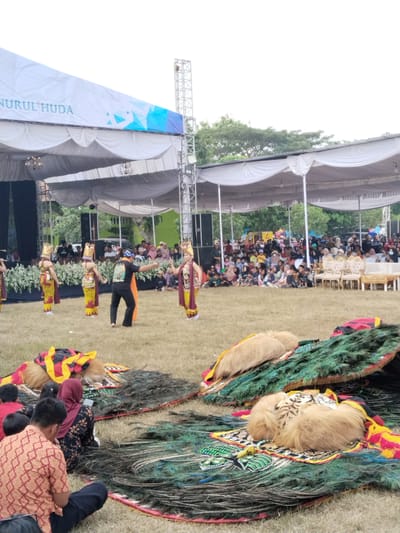 In Plosorejo, Indonesia, an outdoor performance is underway beneath a white tent structure. In the foreground, elaborate costumes featuring peacock feathers and furry accents are laid out on the grass. Several performers in traditional attire, including headdresses, are on stage in the middle ground, engaged in a dance. A person in a black t-shirt and orange fabric is also on stage, possibly directing the performance. A large audience is seated on the grass and on chairs in the background, watching the event. The scene is set against a backdrop of trees and a bright, overcast sky. A banner reading "NURUL HUDA" is visible on the tent.
