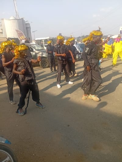 The image depicts an outdoor daytime scene at Itakpe Junction, Nigeria, featuring a procession of approximately 10-15 individuals on an unpaved road. The majority of the people in the foreground and midground are young males wearing matching black shirts, black trousers, and elaborate headwear constructed from golden tinsel or streamers. Their footwear includes various sneakers, such as blue and white and tan and white. One individual in the foreground has a dark strap hanging from his left leg. Another person on the right side of the main group wears a black top with a white number "7" visible on the sleeve. In the background, to the right, several individuals are dressed in bright yellow full-body costumes.

The participants are in motion, some with arms raised or bent, suggesting dancing or a coordinated parade. Long, distinct shadows are cast by the individuals, indicating a low sun angle and clear weather. A dark-colored pickup truck or SUV with a visible dent in the hood is parked or moving slowly behind the group on the left. Industrial-style cylindrical silos or storage tanks are visible in the far-left background, alongside other indistinct buildings and structures. The front wheel and partial frame of a silver motorcycle are visible in the bottom-left corner of the frame. The sky is bright and clear.