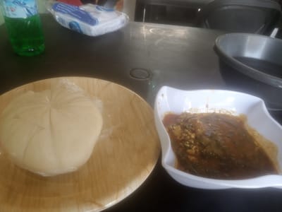 The image shows a tabletop setting in Kinkinau, Nigeria, likely prepared for a meal. The main subjects are two dishes of food. On the left, a mound of what appears to be pounded yam, a staple carbohydrate, is presented on a light brown, circular plate. It's covered in plastic wrap, indicating it might be prepared for takeaway or to keep it fresh. To the right, a white, square bowl contains a rich, dark stew. The stew has a somewhat granular texture and appears to be made with a base of peppers, spices, and possibly meat or fish.
In the background, a green plastic bottle, possibly containing a beverage, stands upright. Next to it is a plastic package of white disposable napkins or tissues, with blue lettering that is partially visible. The table surface is dark and appears to be made of wood or a similar material, with some reflections and faint circular marks suggesting it's a functional surface. There are also glimpses of dark chairs in the background, suggesting a dining area.
The overall scene suggests a casual meal setting, either at home or in a local eatery. The lighting seems to be indoor, and the time of day is indeterminate. There are no people visible in the frame, focusing the attention solely on the food and dining accessories. The mood conveyed is one of simple sustenance and perhaps preparation for consumption. Visible text includes branding on the beverage bottle (partially obscured) and the packaging of the napkins, which reads "AMBISTANTAL CENTERS" and "TEL: 08074789053".