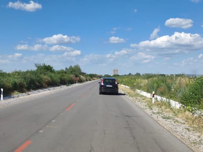This image captures a view of a paved road in Krushovitsa, Bulgaria, on a bright, sunny day with scattered white clouds in a clear blue sky. The road is empty except for a black SUV driving away from the viewer in the middle of the frame. The SUV has a license plate with visible text, although it is not entirely legible.

The sides of the road are lined with lush green vegetation and some dry grasses, suggesting a rural or semi-rural setting. There are concrete barriers and gravel shoulders along the road. Road markers, a white post with red and white reflective strips, are visible on the right side of the road. A distant, tall, light-colored building can be seen on the horizon, possibly an apartment building or hotel.

The scene appears to be set during the daytime, likely late morning or early afternoon, given the strong sunlight casting shadows. The weather is clear and pleasant. The overall impression is one of a peaceful, open road journey in a natural landscape. The red lines painted on the road indicate lane markers or possibly cautionary markings.