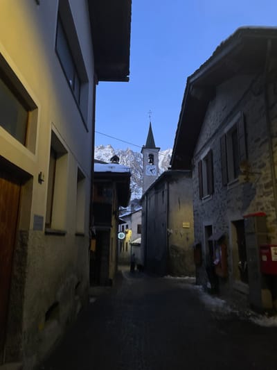 The image, when properly oriented, captures a narrow, atmospheric alleyway in the alpine town of Courmayeur, Italy, during the "blue hour" – either dawn or dusk. Buildings of varying heights and materials, including stone and light-colored plaster, line both sides of the passage, creating a corridor effect.

The focal point in the distance is a distinct church tower with a steeple, topped by a cross, and featuring a clock face. Behind the church, majestic, snow-capped mountains rise under a clear, deep blue sky, indicating an alpine setting. The ground of the alley appears to be paved and shows some patches of snow or ice, suggesting cold weather conditions.

Artificial lighting is visible further down the alley on the right side, casting a subtle glow on the building facades and the ground. One such light is a circular, glowing sign affixed to a building, though any specific text or logo on it is not legible. No people are visible, lending a quiet and tranquil mood to the scene. The architecture and mountain backdrop collectively establish the setting as a historic European mountain village.