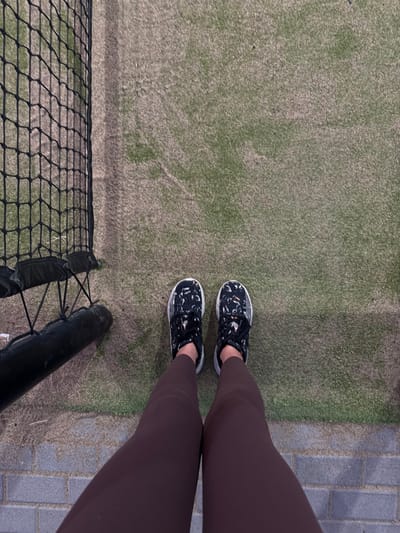 A top-down view shows a person standing on a tennis court wearing dark leggings and patterned sneakers with white soles. The court surface is green and appears slightly worn. A tennis net with a black frame is visible in the bottom left corner. A grey brick pathway borders the court on the right side of the frame.