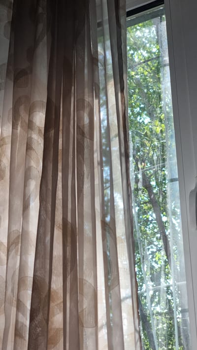 The image shows a close-up of a window with sheer curtains partially covering the view of trees and foliage outside. The curtains are a light brown color with a subtle patterned design of curved lines and circles. Sunlight filters through the sheer fabric, creating areas of light and shadow on the curtain. Through the gaps in the curtains, a green canopy of leaves and branches is visible, suggesting an outdoor natural environment. The window frame appears to be white. There are no people or specific landmarks that would indicate a location in Hakkulabad, Uzbekistan.