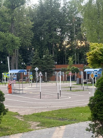 The image captures a driver training course located in the city of Dedovsk, Russia, likely on a paved lot surrounded by trees. The scene features a variety of traffic signs, including yield signs, stop signs, pedestrian crossing signs, and a railroad crossing sign. A small blue shelter with white posts is visible in the mid-ground, and further back, a building with a wooden facade stands behind a row of trees. Traffic lights are also present, along with traffic cones. White lines mark out driving lanes and parking spots on the asphalt surface. The environment appears to be a park or outdoor recreational area, given the lush greenery and mature trees that frame the scene. The sky is overcast, suggesting a cloudy day. There are no people actively engaged in training activities visible in this particular frame.