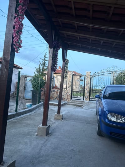The image shows a view from under a wooden structure decorated with artificial flowers. In the background, there is a gate with ornate metalwork, beyond which is a blue car parked on a concrete surface. Further back, a house with stone and decorative elements is visible. A fence runs along the right side of the image, partially obscuring another building. The sky is visible through the gaps in the structures. The location is in Vinitsa, Bulgaria, based on identifying factors from the location.