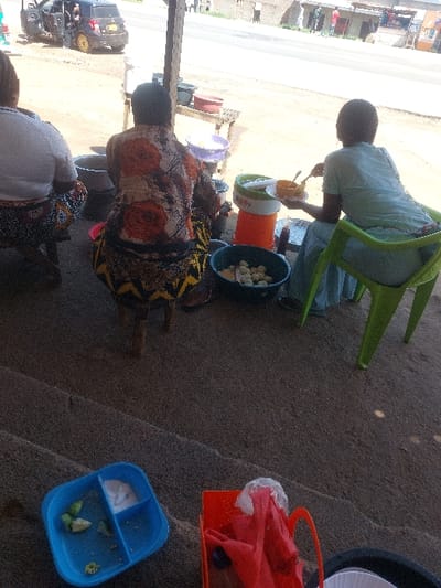 The image depicts an outdoor or semi-outdoor food vending scene in Kimamba, Tanzania, adjacent to a paved road. Three individuals are visible, primarily from their backs.
In the foreground, a blue plastic divided tray contains small green food items and a white powdery substance. Adjacent to it, an orange plastic basket holds red fabric. The ground consists of coarse dirt or concrete.
Centered in the midground, a person wearing a multi-patterned shirt with orange, brown, and black motifs, and a patterned skirt, is seated on a small wooden stool. Large metal pots are positioned in front of this individual, suggesting food preparation. To their immediate right, an orange insulated beverage dispenser and a white plastic plate are visible. A green plastic bucket on the ground contains numerous small, round, light-colored food items.
To the right, a person in a light-colored top and skirt is seated on a lime green plastic chair, holding a small orange bowl and a utensil, appearing to be eating. To the left, a third person is partially visible, wearing a white top and a patterned skirt, seated on a low stool.
In the background, beyond a visible wooden support beam, a paved road is present with a dark-colored SUV-type vehicle parked on the dirt shoulder, its doors open. Distant buildings line the road, and several pedestrians are visible. The overall scene indicates an active roadside food stall or informal eatery.
