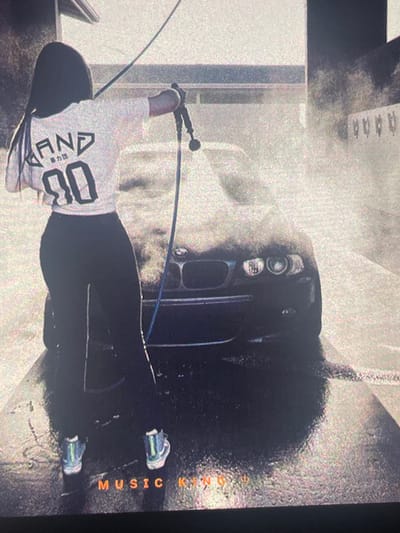 The image depicts a woman washing a black car at a car wash. She is wearing a white t-shirt with Japanese characters and the number "90" on the back, and black leggings. She is holding a high-pressure washer and spraying water on the car. The car wash appears to be an outdoor facility with a concrete floor. The background shows a building structure and some white railings. The image also has text at the bottom that reads "MUSIC KINGDOM". The scene is likely set in Paczkowo, Poland, given the context.