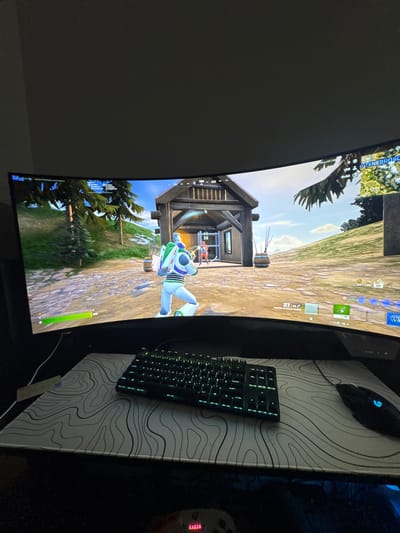 A curved computer monitor displays a Fortnite game with a Buzz Lightyear character running towards a wooden structure. A black keyboard with illuminated keys and a black gaming mouse sit on a mousepad featuring a white topographical map pattern. The monitor, keyboard, and mouse are situated on a dark surface. The location is Glendale Heights, United States of America.