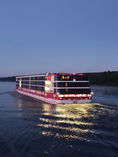 A panoramic, eye-level, medium shot captures a modern, multi-decked cruise ship, painted in white and striking pink, navigating on a calm body of water at dusk. The ship, identified by Russian text on its hull, displays illuminated windows across its numerous decks, casting a warm, golden glow onto the dark, rippling water. The vessel is creating a visible wake behind it, with white foam churning in its path. The background features a dense, dark treeline rising from the far bank of the water, silhouetted against a fading twilight sky that transitions from a deep blue overhead to a lighter hue near the horizon. The overall scene suggests a serene moment of travel and illumination in a natural, tranquil setting. The text on the ship reads "МУСТАЙ КАРИМ" and "САНКТ-ПЕТЕРБУРГ".