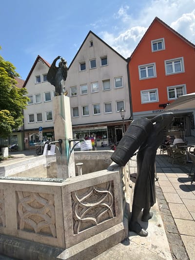 The image depicts a public square in Ebermannstadt, Germany, featuring a bronze sculpture of a person carrying a large barrel. The sculpture is located in front of a fountain with a decorative stone basin. In the background, there are several buildings, including a prominent orange-colored structure and a light-colored, multi-story building with numerous windows. A lamppost stands near the orange building. An awning is extended over a cafe or restaurant area with outdoor seating, where two people are visible sitting at a table. The ground is paved with cobblestones. A tree with green foliage is visible on the left side of the frame. The sky is blue with scattered white clouds. The scene appears to be from a sunny day.