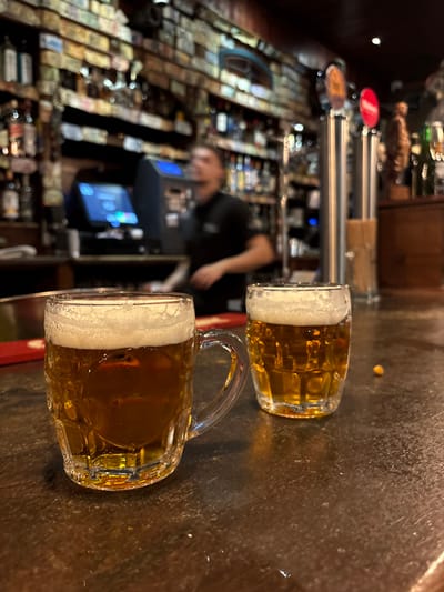The image depicts an indoor bar scene in the city of Cádiz, Spain. In the immediate foreground, two clear glass mugs filled with golden-amber beer and topped with white foam rest on a dark, worn bar counter. The mug on the left is a handled tankard, while the one on the right is a smaller, handle-less glass. A single yellow popcorn kernel is visible on the counter to the right of the smaller mug. Behind the mugs, a red trim lines the bar's edge.

In the midground, a male bartender, blurred due to motion, is visible behind the counter, wearing a dark shirt and facing right. A point-of-sale system with a blue-lit screen is situated to the bartender's left. To the right, two metallic beer taps, one with an orange label and another with a red label, are prominent. The background features extensive shelving units stocked with numerous bottles of various spirits. The wall above these shelves is covered in a dense mosaic of small, rectangular paper items, likely currency notes or labels. The overall lighting is dim, consistent with an evening bar environment.