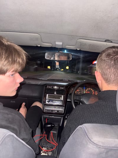 Two young men are inside a car at night, with the view from the passenger seat looking towards the driver. The passenger in the foreground, with light brown hair, is looking to his left, holding a black smartphone in his right hand. The driver, also with light brown hair, is wearing a dark grey knitted sweater and is focused on the road ahead, with his hands on the steering wheel. The car's dashboard is visible, showing a stereo system, gear shifter, and car instruments. The road outside the car is illuminated by streetlights and appears to be in an urban or suburban area. The environment suggests a journey taking place on a paved road within a populated area. There is no indication of the specific location within Ptichnik, Russia.