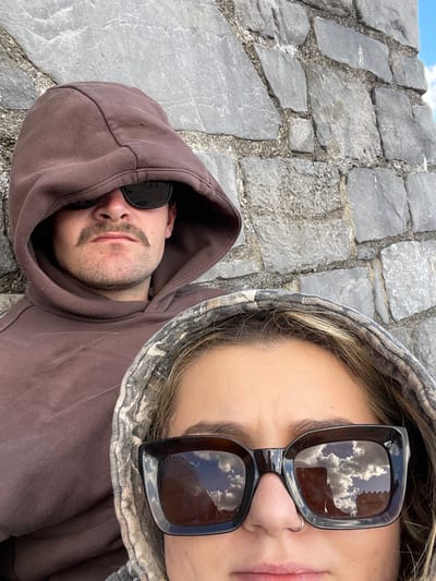 The image features two individuals positioned in a close-up, slightly angled shot, against a stone wall consistent with structures found in Tenby, United Kingdom. The background consists of a grey, irregularly coursed stone wall with visible mortar. A sliver of blue sky with white clouds is discernible in the upper right corner and reflected in one individual's sunglasses.
On the left, an individual wears a solid brown hooded sweatshirt, with the hood pulled up. Dark sunglasses obscure the eyes, and a dark mustache is visible on the upper lip.
In the foreground, on the right, another individual wears a camouflage-patterned hooded sweatshirt with the hood pulled up. Large, dark, rectangular sunglasses cover the eyes, reflecting a cloudy sky and faint architectural elements. A small silver ring is present in the left nostril. Both individuals are oriented towards the camera.