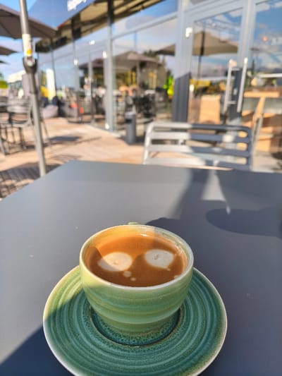 The image foregrounds a light green ceramic cup filled with coffee, topped with crema exhibiting two distinct light circular patterns, resting on a matching ridged saucer. These items are positioned on a dark grey outdoor table, with sunlight casting shadows from the left. The blurred background reveals an outdoor patio with a wooden deck, furnished with multiple dark tables, chairs, and open dark patio umbrellas. Behind this, a modern building with extensive glass windows and light-colored frames reflects portions of the outdoor setting and sky. A light grey slatted bench is visible near the building. No identifiable individuals or specific actions are discernible. The scene is an outdoor café or restaurant area in Hermsdorf, Germany, under bright conditions.