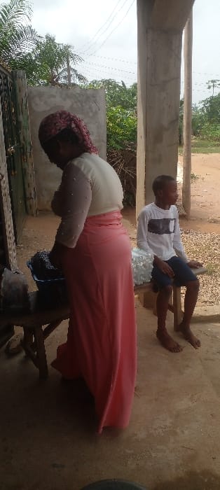 The scene depicts two individuals in an outdoor area within Agunmo, Nigeria, framed by building structures and natural elements. An adult female stands centrally, viewed from the back-left, wearing a patterned red, black, and white head covering, a light-colored long-sleeved top with pearl-like embellishments on the sleeves, and a flowing pink maxi skirt. She is leaning over a blue plastic basin filled with dark, lumpy items, which rests on a wooden stool. To her right, a young male, appearing to be a pre-teen, sits barefoot on a second wooden stool. He wears a white long-sleeved shirt with a black graphic design and dark shorts, his gaze directed to the right, off-camera. A cluster of clear plastic bags, possibly containing liquid, is positioned on the stool beside him. The ground consists of light brown, unpaved earth or concrete. The background features dense green vegetation, including palm trees, utility poles, and overhead power lines against an overcast sky. A dark metal gate or door is partially visible on the far left, while an unrendered concrete pillar frames the right side of the immediate foreground. No direct interaction between the individuals is observed.