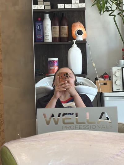 Here is a detailed description of the image:
**General Scene:**
The image appears to be a selfie taken in a hair salon, possibly in Arroyo de la Miel-Benalmádena Costa, Spain. The reflection in the mirror shows a person sitting in a hair salon chair, getting their hair washed in a salon sink.
**People:**
* There is a person visible in the mirror's reflection holding up a cell phone and likely taking the picture. Only the person's head and hands are fully visible.
**Objects:**
* **Hair Salon Sink:** A white, ceramic hair salon sink is present, where the person is having their hair washed.
* **Hair Products:** Shelves with various hair products, including shampoo and styling products, are visible behind the sink. Some product names/brands can be partially read, such as "Kolex" and "PEstiVio."
* **Mirror:** The scene is viewed through a large mirror with a logo.
* **Logo:** A logo for "Wella Professionals" is partially visible on the mirror's right side.
* **Cell Phone:** The person is holding a gold-colored cell phone (likely an iPhone) to take the selfie.
* **Light Fixture:** An orange salt rock with a white camera light is placed on the top shelf.
**Environment:**
* **Hair Salon:** The environment is a typical hair salon setting, with hair washing equipment and product displays. The location of the salon may be in Arroyo de la Miel-Benalmádena Costa, Spain.
**Action:**
* A person is getting their hair washed in the salon.
* The person is taking a selfie in the mirror.