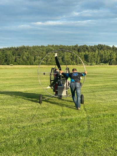 A person is pushing a powered parachute on a grassy field under a cloudy sky. The person, viewed from behind, is wearing blue jeans and a long-sleeved shirt with a blue and white design. They are holding onto the frame of the parachute, which has a large propeller at its center and two wheels. The parachute appears to be a paramotor. The background features a line of trees and a vast expanse of green grass. The scene is illuminated by natural light, suggesting it is daytime. The location is identified as Турово, Russia.