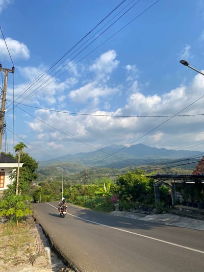 The image captures a view of a rural road winding through a verdant landscape, likely in the city of Kresek, Indonesia. A paved road with a white line down the center curves to the left in the foreground, leading towards rolling hills and distant mountains under a bright blue sky dotted with fluffy white clouds.

On the road, a motorcycle with two occupants, a driver and a passenger, is traveling away from the camera. Both individuals are wearing helmets.

To the left of the road, a white house with a dark roof and a prominent wooden-framed window is visible. Next to the house, a utility pole stands tall, laden with numerous electrical wires that stretch across the frame, some of which are silhouetted against the sky. Lush green vegetation, including what appears to be a papaya tree, grows along the roadside. A small, eroded ditch with exposed rebar runs alongside the edge of the road.

To the right of the road, a property is visible with a covered entrance featuring an arched gateway, stone pillars, and various potted plants. A streetlamp is positioned along the roadside, pointing upwards. The overall environment is characterized by abundant greenery, characteristic of a tropical or subtropical region. The lighting suggests it is daytime, with sunlight casting shadows on the road.