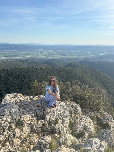A woman sits on a rocky cliff edge, gazing towards the camera, with a vast panoramic vista stretching out behind her. She wears a light blue shirt, light-colored pants, sunglasses, and red shoes. The background features a wide valley or plain with green fields and distant hills, under a blue sky with visible contrails. The rocky terrain around her is interspersed with sparse vegetation. A small yellow object resembling a bottle is on the rock next to her. The scene suggests a natural landscape, possibly a mountaintop location. The city of Bonnieux, France is possibly visible in the distant valley.
