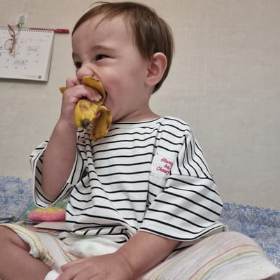 The media file shows a close-up, rotated shot of a young child with brown hair eating a banana. The child is wearing a black and white striped t-shirt that has a small red embroidered message on the left chest area that reads "Always be Cheery". The child's face is partially obscured by the banana they are eating. Their left ear is visible, and their dark eyes are focused on the banana. The child is holding the banana with their left hand, with the peel still attached.
The background appears to be indoors, with a textured, light-colored wall on the left. A blue and white patterned fabric, possibly a bedspread or blanket, is visible in the upper right portion of the image. On a surface below the child, a notebook or planner with some handwritten notes and diagrams is visible. A pair of glasses rests on top of the notebook, and a red pen is placed to the left of it. The lighting suggests it is daytime, with natural light illuminating the scene. There are no clear indications of the weather. The image conveys a sense of a casual, everyday moment.