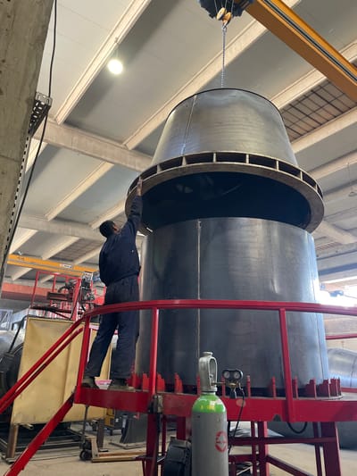 An individual wearing dark clothing is positioned between a large metal cylinder suspended by a yellow crane and a red metal frame or scaffold. The cylinder has a flared end with internal structures. The person appears to be reaching towards the cylinder. The red frame has a green and silver gas tank attached to it. The location appears to be an industrial building, possibly a factory or workshop, with a high ceiling and structural supports visible. Tomba, Montenegro is not identifiable in the image.