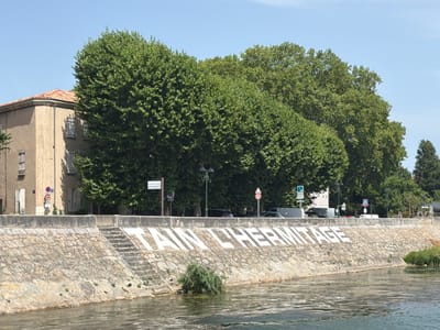 The image shows a stone embankment alongside a body of water. Large white lettering is painted on the embankment, spelling out "TAIN L'HERMITAGE". The embankment is made of rough-hewn stones and has a set of stairs leading up from the water's edge. A dense line of trees forms the background, with a building visible on the left side of the frame. Several streetlights and traffic signs are also present along the top of the embankment. The sky is clear and blue. The location is identified as Mercurol-Veaunes, France.