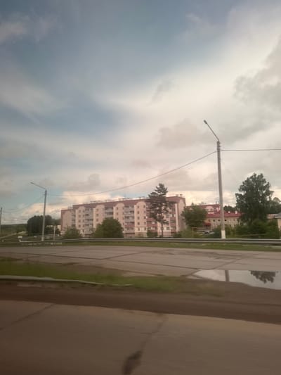 The image depicts a static urban landscape in Kizner, Russia, under an overcast sky, suggesting cloudy weather or recent rain. The photo appears to be taken from a slightly elevated perspective, possibly from inside a vehicle, looking towards the left.

The sky occupies a significant portion of the upper and left side of the frame, filled with heavy, grey clouds, indicating diffused light characteristic of an overcast day.

In the mid-ground, a row of multi-story residential buildings, likely apartment blocks, lines what appears to be a street. These buildings feature light-colored facades, possibly beige or off-white, and have distinctive reddish-brown sloped roofs. Trees with green foliage are interspersed among the buildings, providing a touch of nature to the urban environment. Tall utility poles with streetlights and overhead wires are prominent, running along the street parallel to the buildings.

The foreground and right side of the image feature a wide, paved surface, which appears wet and reflective, strongly suggesting that it has recently rained. This surface might be a road, a large sidewalk, or a plaza. A thin strip of green grass or dirt separates this wet pavement from the road where the utility poles are located. No specific activity is visible; the scene is calm and devoid of people or moving vehicles. There is no visible text in the image.