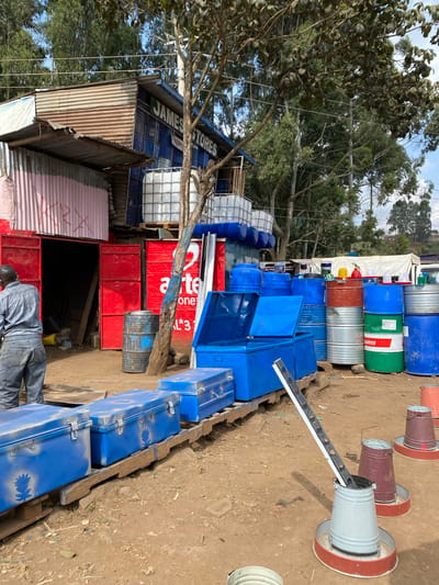 The image depicts an outdoor market scene in Limuru, Kenya. In the foreground, several large blue metal trunks with latches are arranged on wooden pallets. To the right of the trunks, there are several cylindrical chicken feeders, some metal and some plastic, with red bases. In the midground, a variety of blue plastic containers and barrels are stacked. A red-painted metal barrel is positioned near a tree. Behind this, a building with a corrugated metal roof is visible, with red double doors open. A sign on the building reads "JAMES STORES". To the right of the building, a large red sign with "a te" and a phone number is visible. In the background, more large white water tanks are stacked, and lush green trees and power lines are present against a partly cloudy sky. A man in a grey shirt and blue jeans is standing facing away from the camera, appearing to work in the market.
