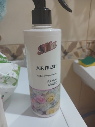 This image shows a bottle of "Sila Air Fresh" floral magic air freshener. The bottle is white with a black spray nozzle. The label features the "Sila" logo, the text "AIR FRESH," and "floral magic." Below "AIR FRESH" in smaller text, it reads "парфум для приміщень" which translates to "perfume for premises." The lower part of the bottle has a floral design.
The bottle is placed on a white surface, possibly a countertop or shelf. To the left, there is a partially visible white package with green accents and floral patterns, suggesting it might be a tissue box or another household product. In the background, a tiled wall with neutral-toned tiles is visible, and to the right, a white bathtub with a yellow and green sponge is present. The overall environment appears to be a bathroom or a similar domestic setting.
The image does not provide any information about the city of Grigoriopol, Moldova.