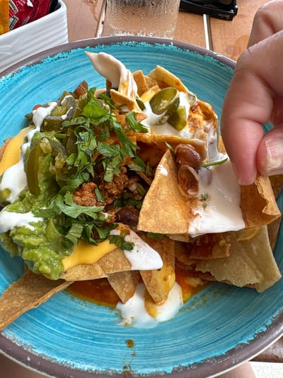 This is a close-up, overhead shot of a plate of nachos, likely taken outdoors in daylight, given the lighting and the presence of sunglasses on the left side of the frame. A person's hand is visible, reaching down to pick up a chip from the plate. The nachos are served in a rustic, blue-rimmed bowl and are topped with a generous amount of cheese sauce, ground meat, jalapeños, guacamole, and a dollop of sour cream or crema. The toppings are vibrant and fresh, suggesting the dish is either newly served or recently prepared. The setting appears to be casual, possibly a restaurant patio or an outdoor dining area, with a wooden surface visible beneath the bowl. The overall impression is one of delicious, hearty food being enjoyed in a relaxed atmosphere. The location context suggests it is Punta Sam, Mexico. There is no visible text in the image.