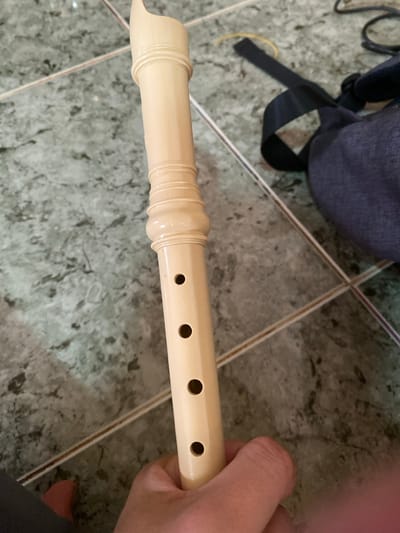 The scene depicts a light-colored recorder held by a person's right hand, positioned vertically against a tiled floor in Đak Đoa, Vietnam. The recorder, appearing cream or light beige, features a visible mouthpiece section, a middle section with two decorative ribbed bands, and a lower section displaying four open circular finger holes. The hand grasps the lower part of the instrument. The floor consists of square, light gray tiles with darker gray patterns, separated by light brown grout lines. In the upper right background, a dark gray fabric bag or backpack is visible, accompanied by a black strap and a thin yellow string or cable. A reflective silver metal rod or pipe extends diagonally across the floor from the upper right to the lower left, passing behind the recorder and the bag. A partial view of a foot or leg, possibly clad in dark fabric, is present in the lower left corner of the frame.