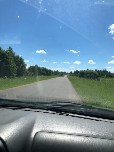 The image, taken from inside a vehicle in Izdeshkovo, Russia, shows a view through the windshield on a clear, sunny day. A grey asphalt road curves into the distance, bordered by vibrant green grass and dense trees on both sides. The sky is a bright blue, dotted with fluffy white clouds. The foreground of the image is dominated by the textured grey dashboard of the car, with the windshield wipers visible at the base of the windshield. There are no people or animals visible in the scene.