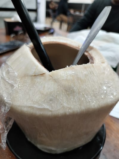A close-up shot shows a whole coconut on a dark plate. The coconut is wrapped in plastic and has a black straw and a silver spoon inserted into its opening. The coconut's flesh is visible inside the opening. The background is blurred, showing a wooden table and hints of other objects and people, suggesting an indoor setting. The image is taken from a high angle, focusing on the coconut.