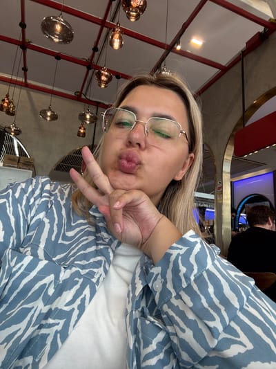 A woman with blonde hair and glasses is taking a selfie in a dimly lit restaurant. She is wearing a blue and white patterned shirt and has her hand on her chin with two fingers extended, making a peace sign. Her lips are pursed, and she is looking at the camera. In the background, there are several hanging lights and arches that suggest an indoor dining setting. The overall ambiance is casual and relaxed.